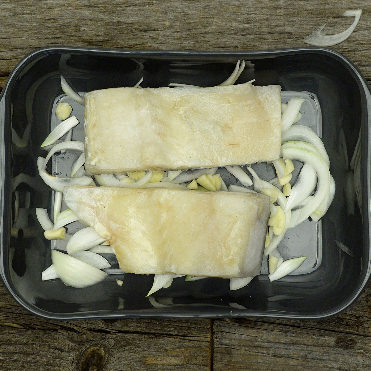 Lombos de bacalhau crus posicionados sobre a cama de cebola e alho na travessa