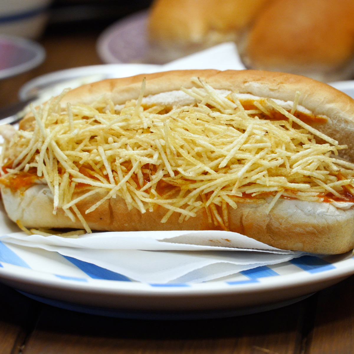 Cachorro-quente sendo montado com molho de salsicha, milho e batata palha.