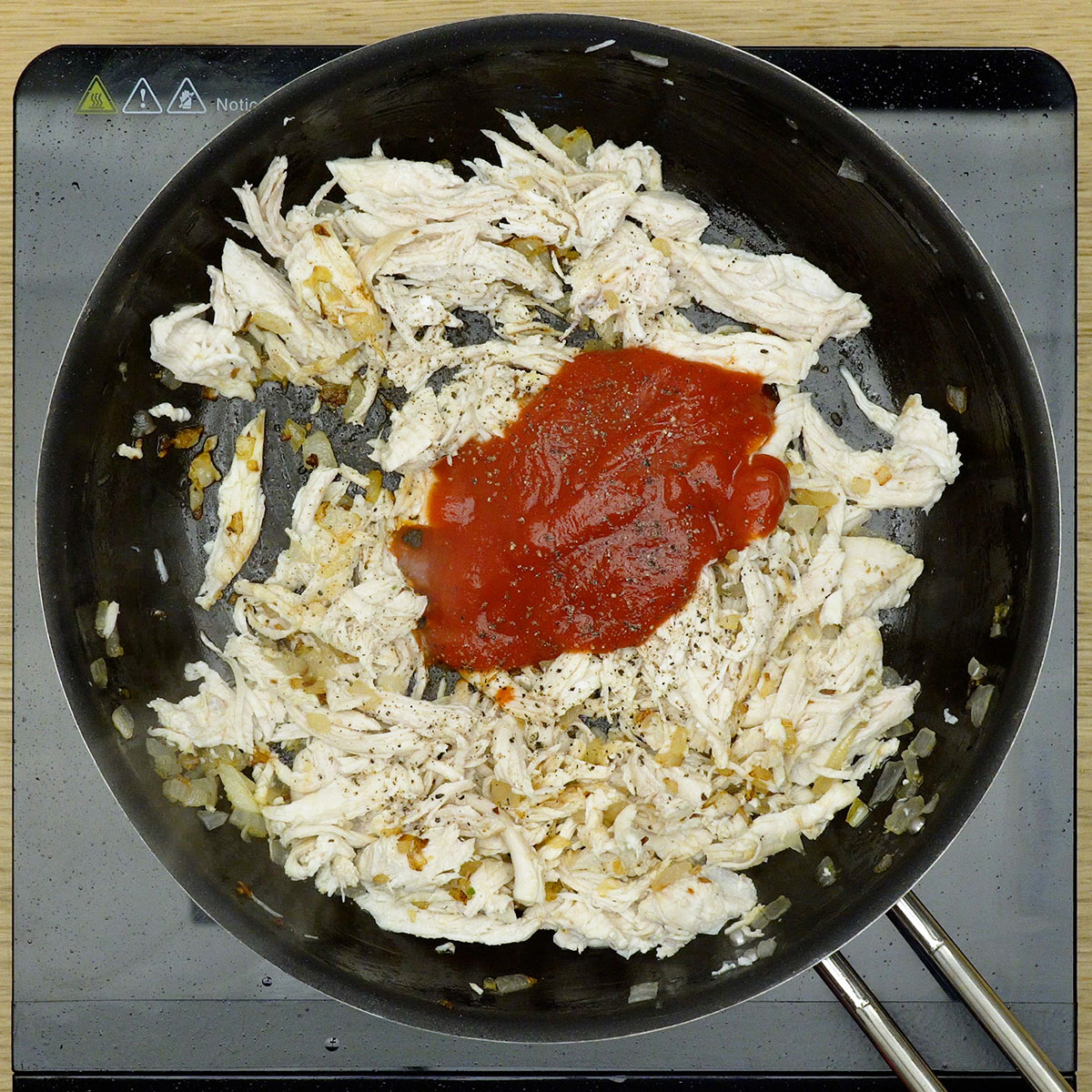 Frango desfiado sendo misturado com temperos e extrato de tomate na frigideira.